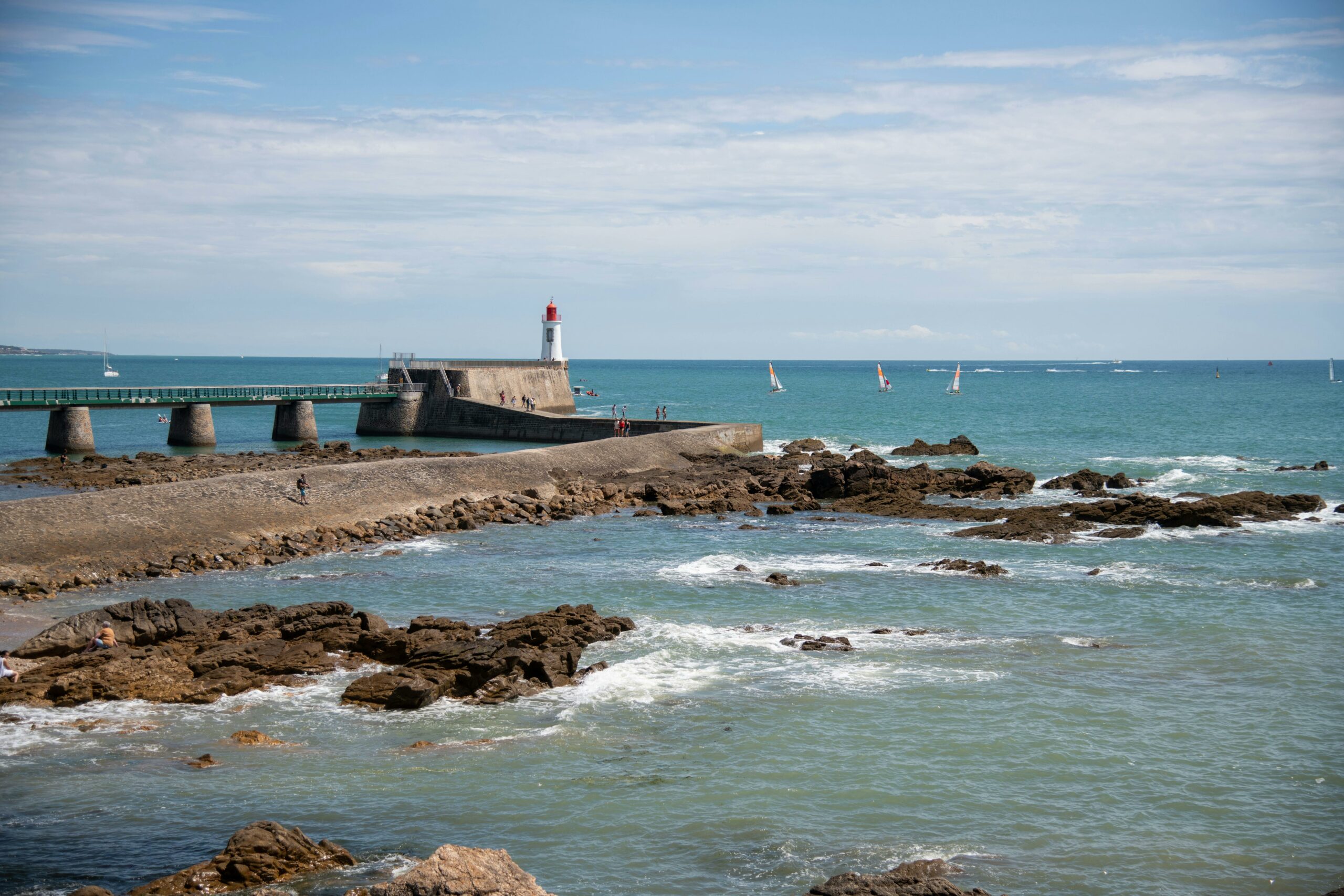 Picturesque view of a lighthouse by the sea with a rocky shore and sailboats on a sunny day.